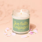 A lit candle in a glass jar with text "Joyfully Engaged VOWED" and pink petals.