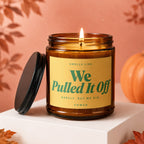 Amber jar candle with lid off and lit flame, reads "Smells like We Pulled It Off".