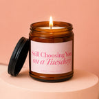 Amber jar candle with lit wick and lid propped open, reads "Still Choosing You on a Tuesday".