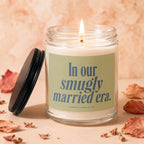 Glass jar candle with lit wick and green label reading "In our smugly married era.".