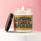 Lit candle in a jar with a black lid, reading "Smells Like Someone Else's Open Bar."