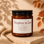 Amber candle labeled "Daughter in Law" with definition text on a stone surface with cream scarf.