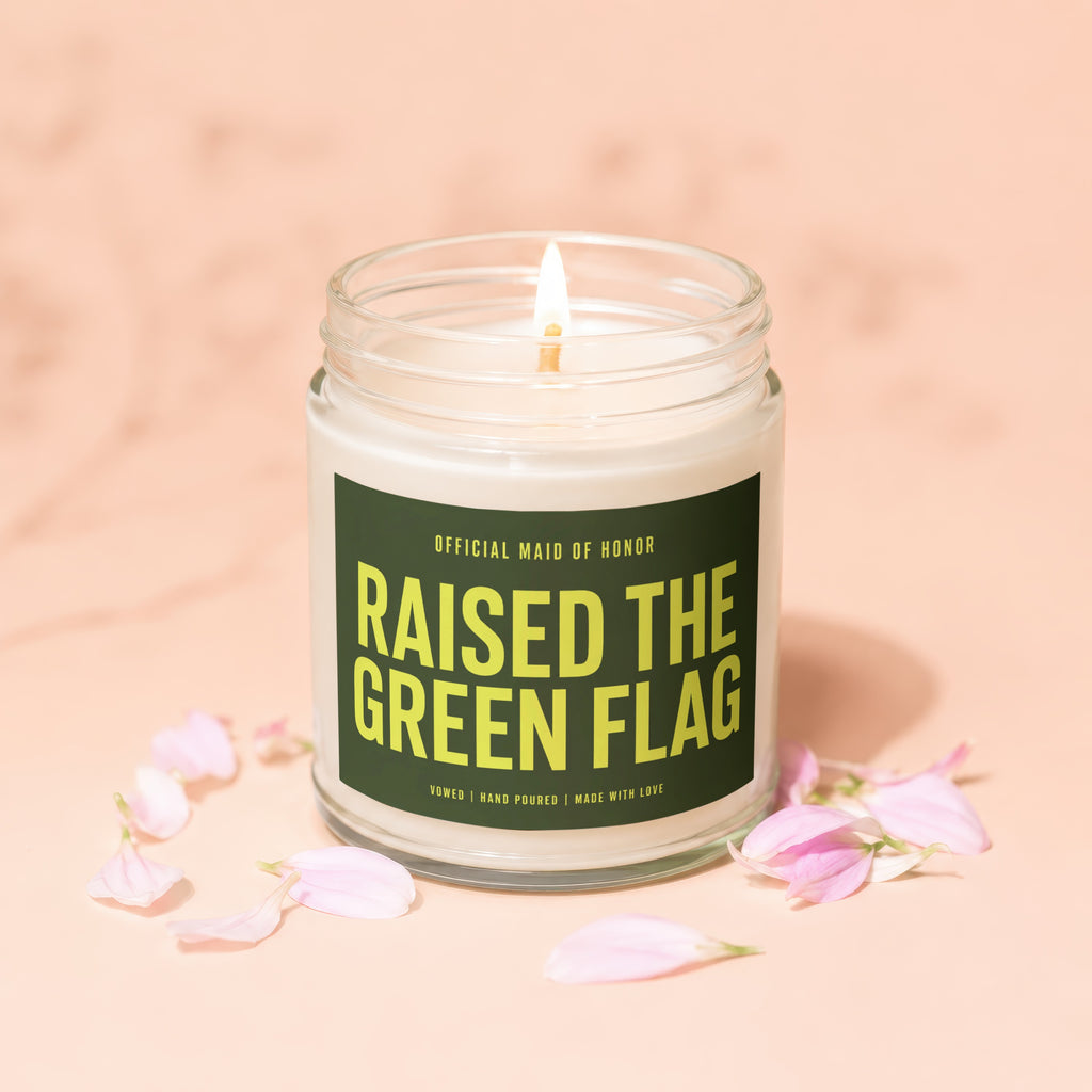 Candle with "Raised the Green Flag" text in a glass jar with pink petal accents.