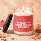 Glass candle with lit wick, red label reads "VOWED LIGHT ME WHEN YOU SAID YES BEFORE YOU SAW THE DRESS".