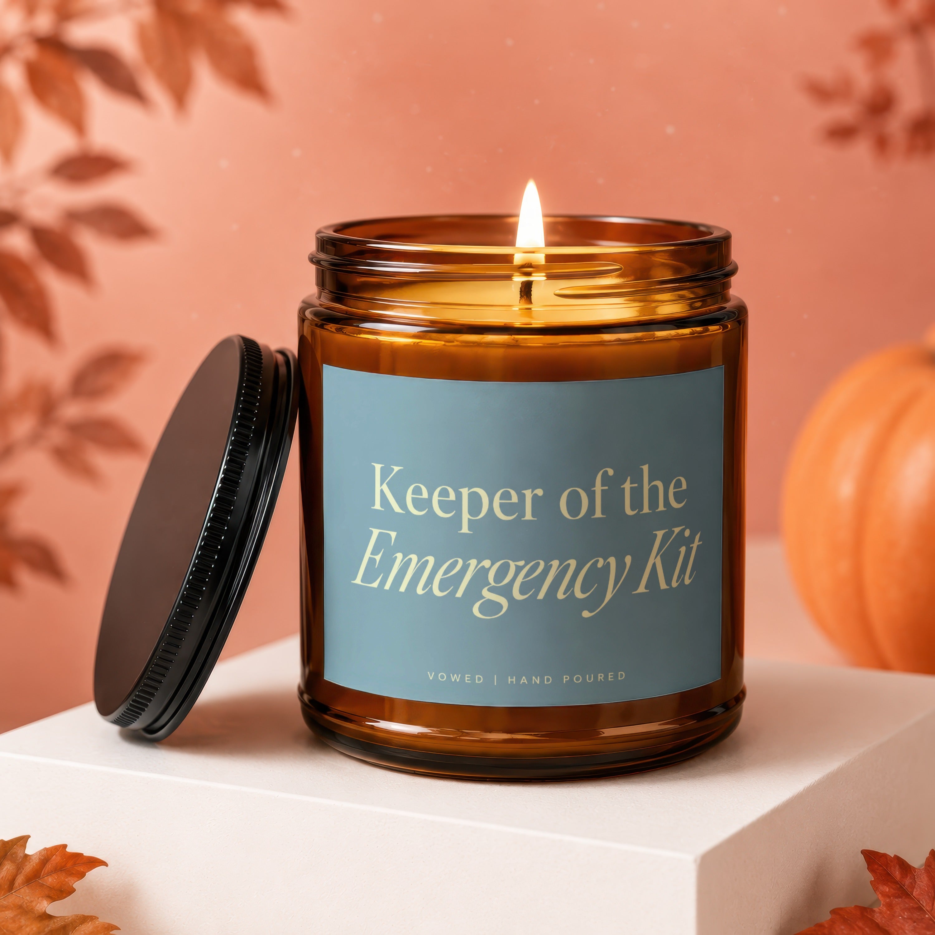 Amber jar candle with a lit wick and a black lid, reading "Keeper of the Emergency Kit."