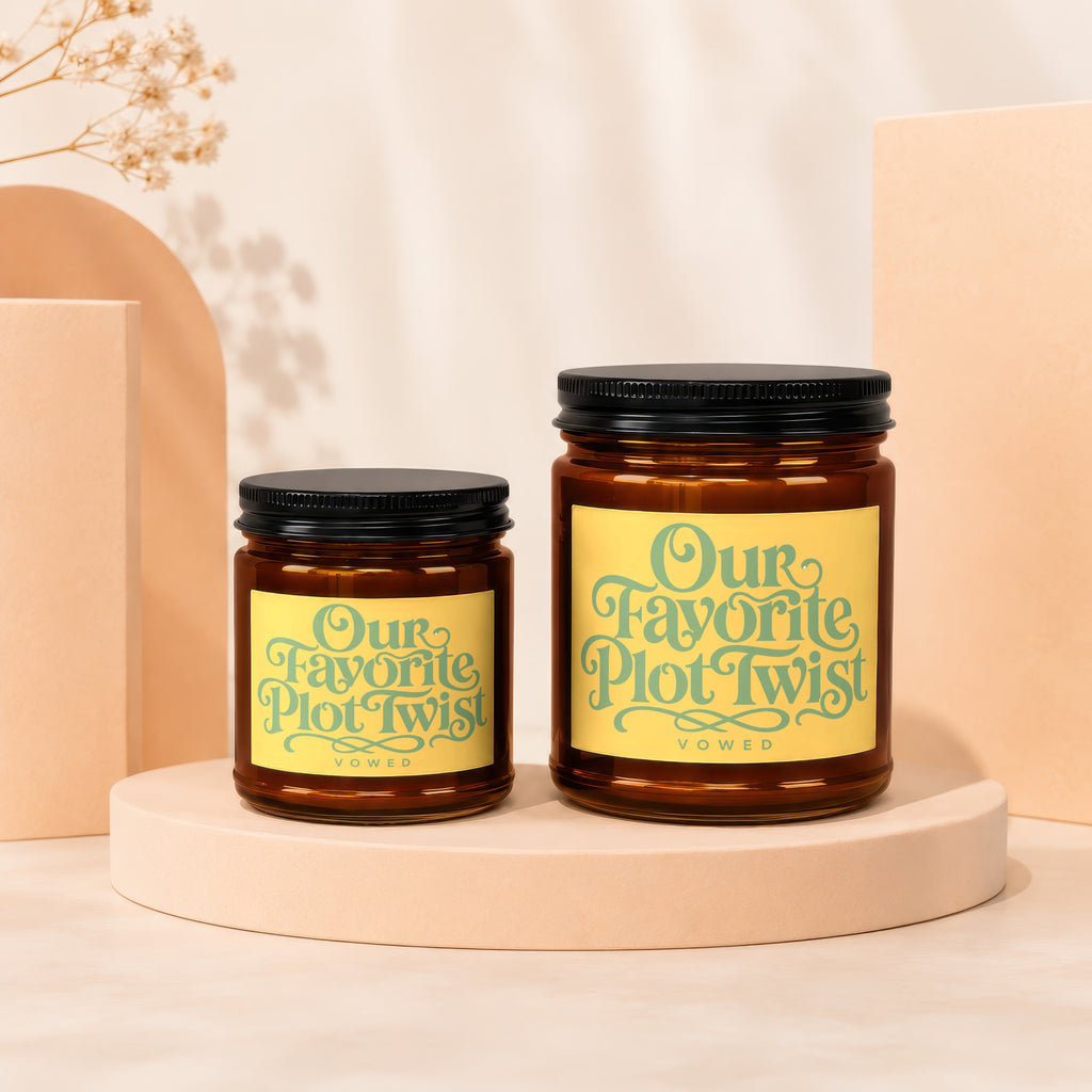 Two amber glass candles with black lids and yellow labels reading "Our Favorite Plot Twist."