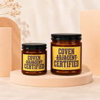 Two amber glass candles with black lids and yellow labels reading "Coven Adjacent Certified".