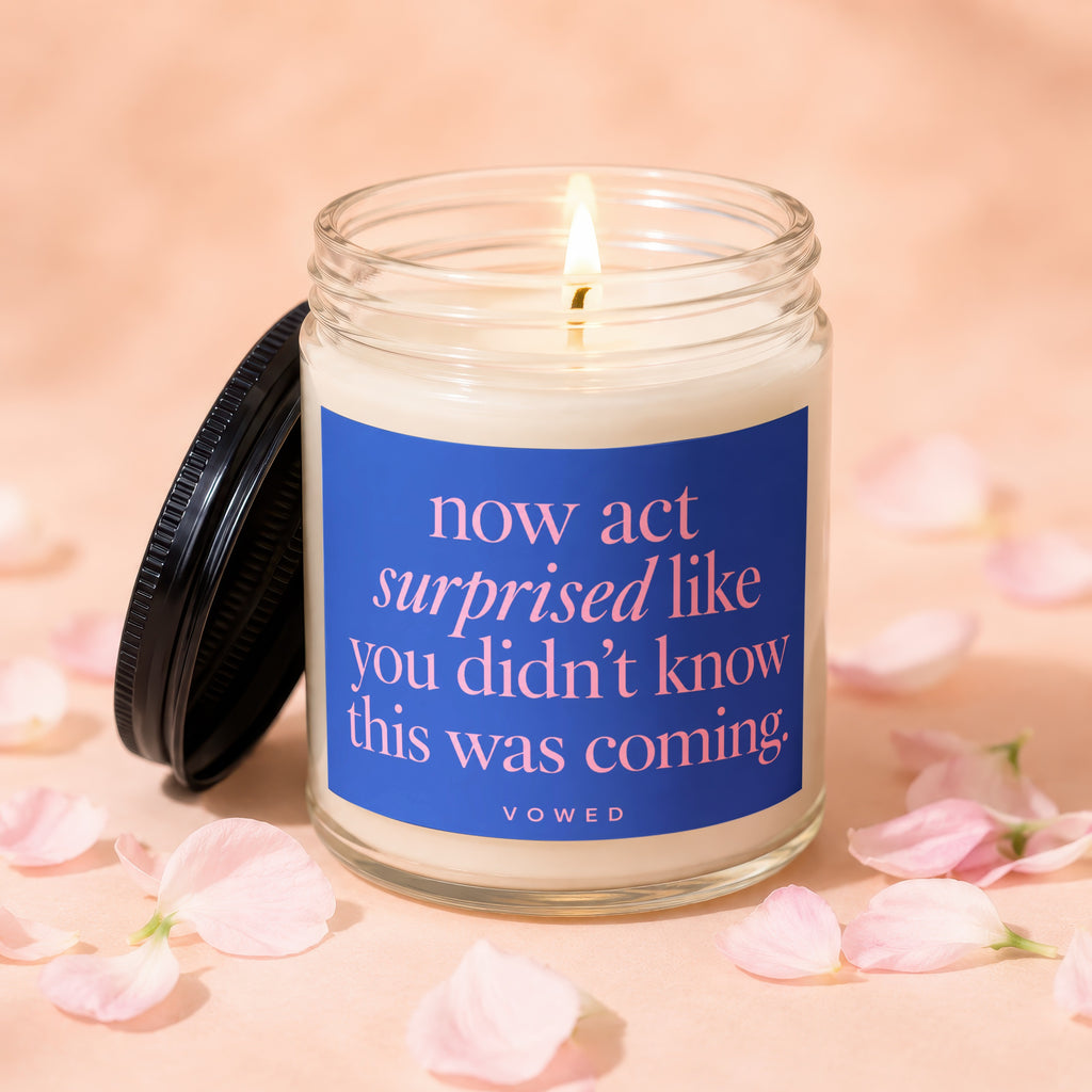Jar candle with lit wick and blue label reading "now act surprised like you didn't know this was coming."