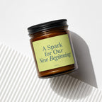 Amber jar candle with black lid, light green label reads "A Spark for Our New Beginning".