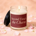 Glass candle with lit flame and burgundy label reading "Second Time's the Charm".