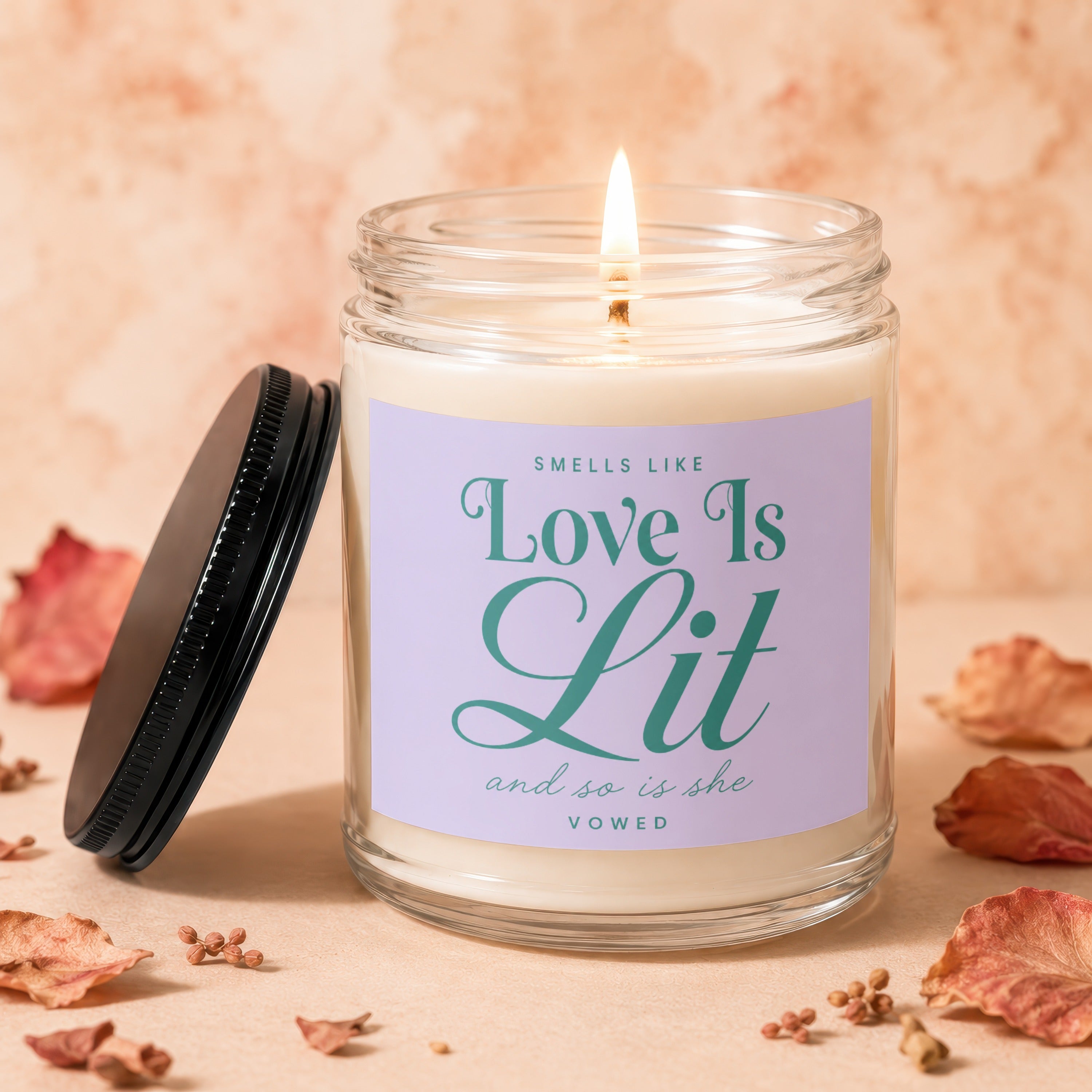 Glass candle with lit wick and lid, reads "Smells Like Love Is Lit and so is she Vowed."