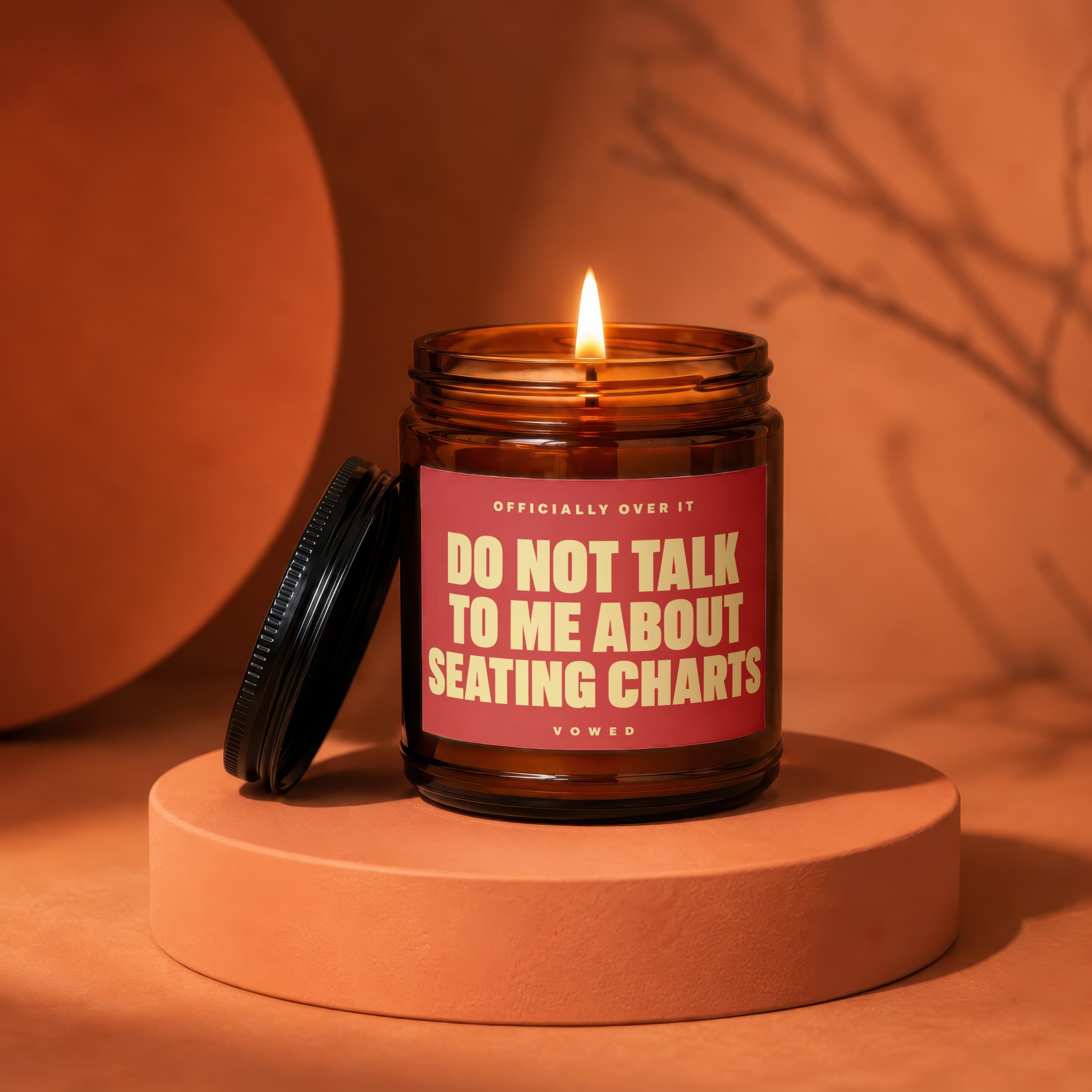 Amber glass candle with lit wick and red label that reads "Do Not Talk To Me About Seating Charts".