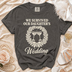 Charcoal gray t-shirt with cream text reading "WE SURVIVED OUR DAUGHTER'S Wedding" and a wreath graphic.