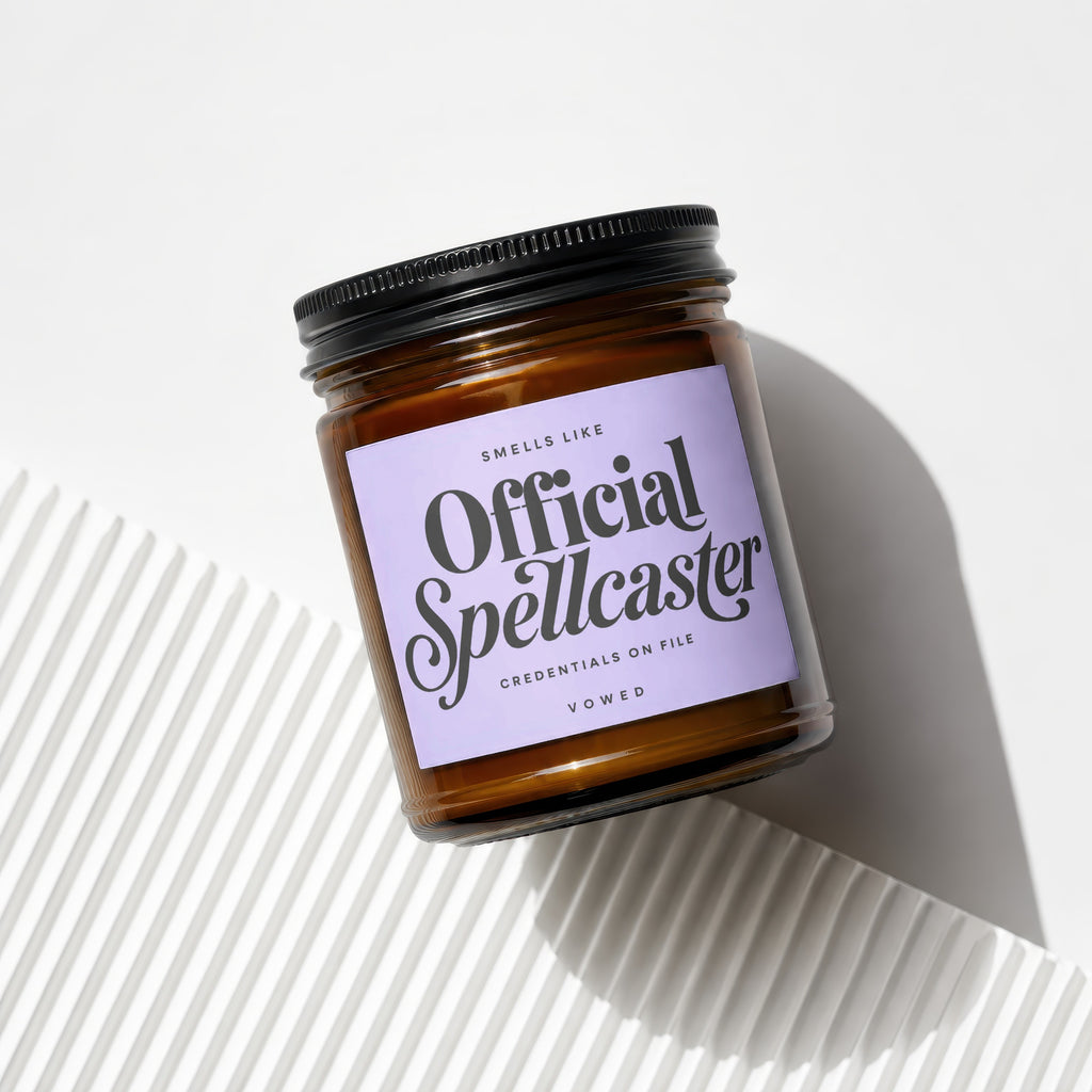 Amber glass jar candle with black lid and lavender label reading "Official Spellcaster."
