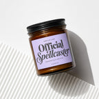 Amber glass jar candle with black lid and lavender label reading "Official Spellcaster."