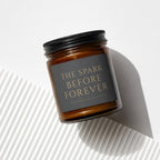 Amber glass candle with black lid and dark gray label reading "THE SPARK BEFORE FOREVER."
