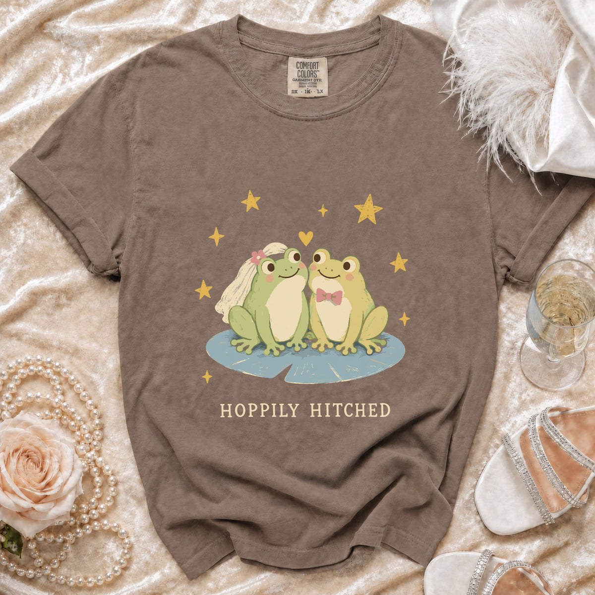 Brown t-shirt with cartoon frogs wearing wedding attire and "Hoppily Hitched" text.