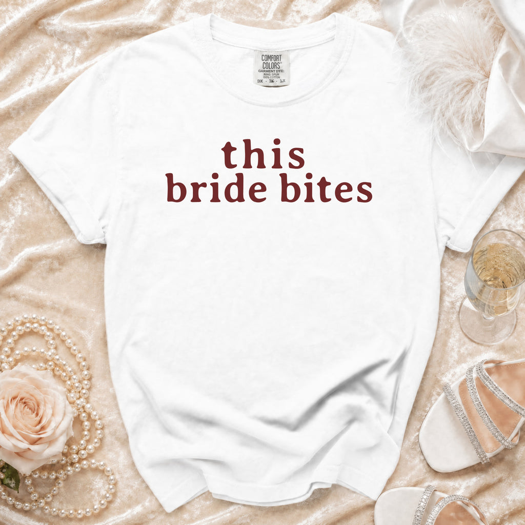 White t-shirt with "this bride bites" text in maroon, laid on velvet with pearls and heels.
