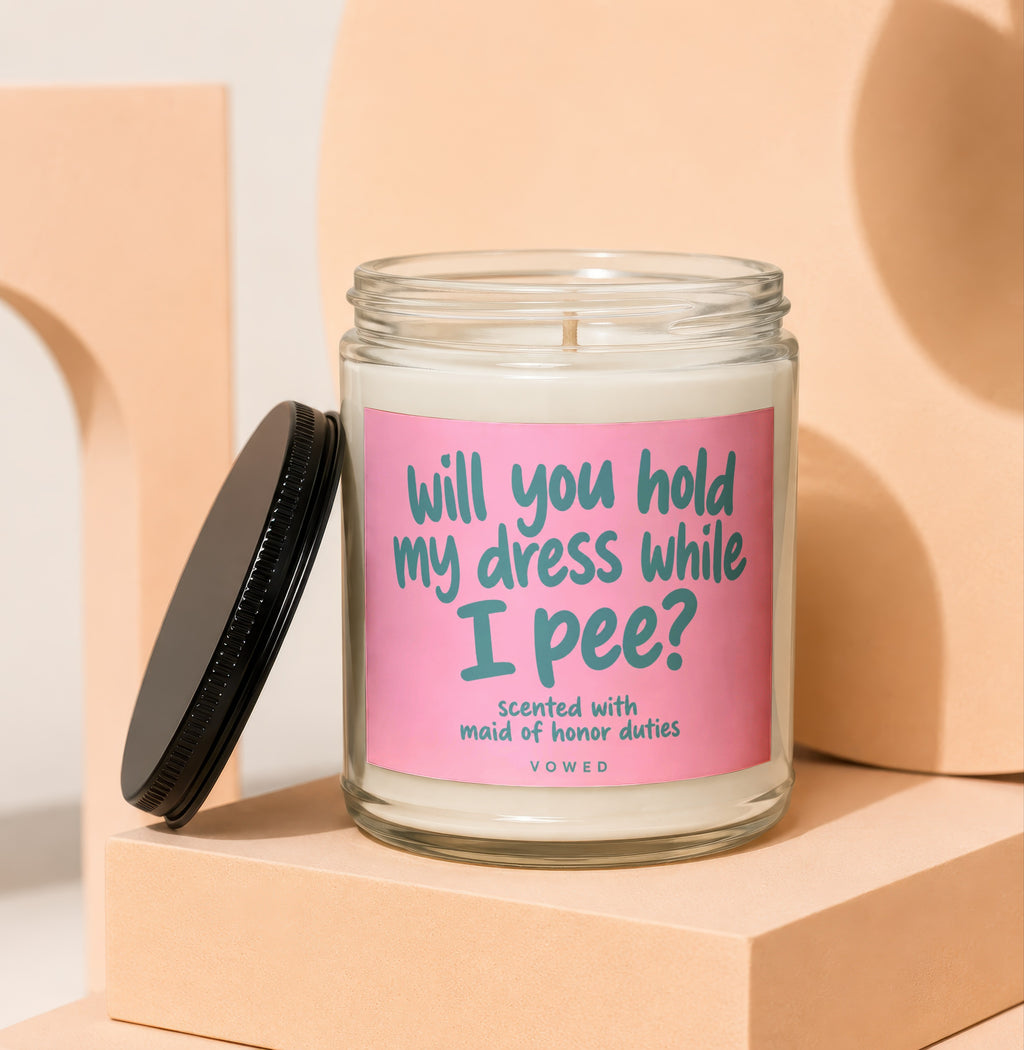Glass candle with pink label reading "will you hold my dress while I pee?"