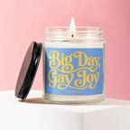 Candle with "Big Day, Gay Joy" text on blue label, lid off, lit wick.