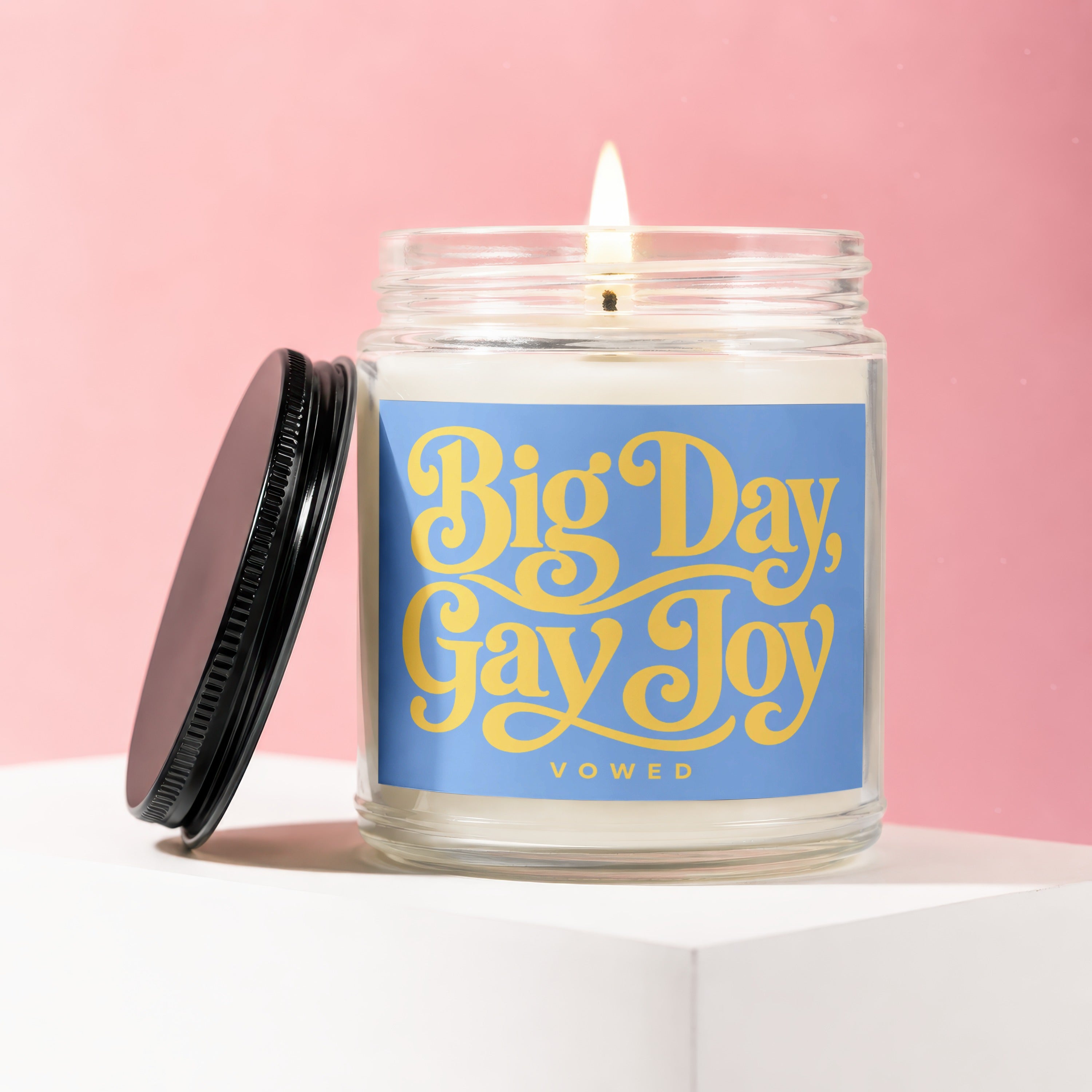 Candle with "Big Day, Gay Joy" text on blue label, lid off, lit wick.
