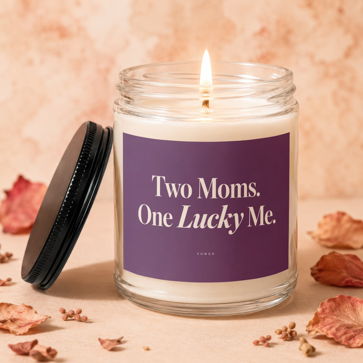 Lit candle in jar with "Two Moms. One Lucky Me." text, lid beside it.