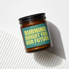 Amber candle with black lid, featuring a teal label with yellow text "STATUS: BURNING BRIGHT FOR OUR FUTURE".