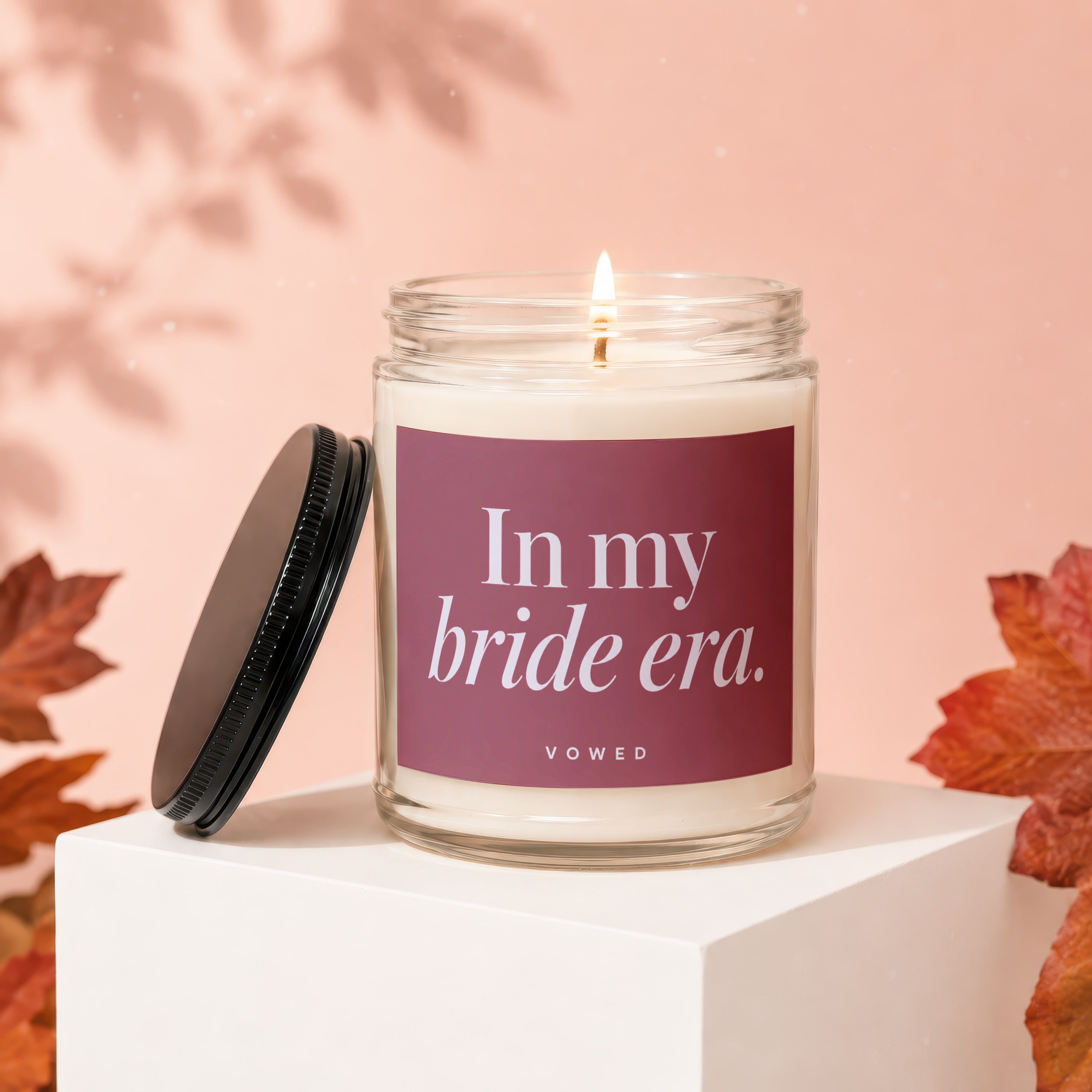 Candle with "In my bride era." text, lid off, on a white cube.