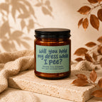 Amber jar candle with black lid reads "Will you hold my dress while I pee?"