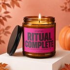 Amber jar candle with lit wick, pink label reads "STATUS: RITUAL COMPLETE ALL PARTICIPANTS ACCOUNTED FOR VOWED".