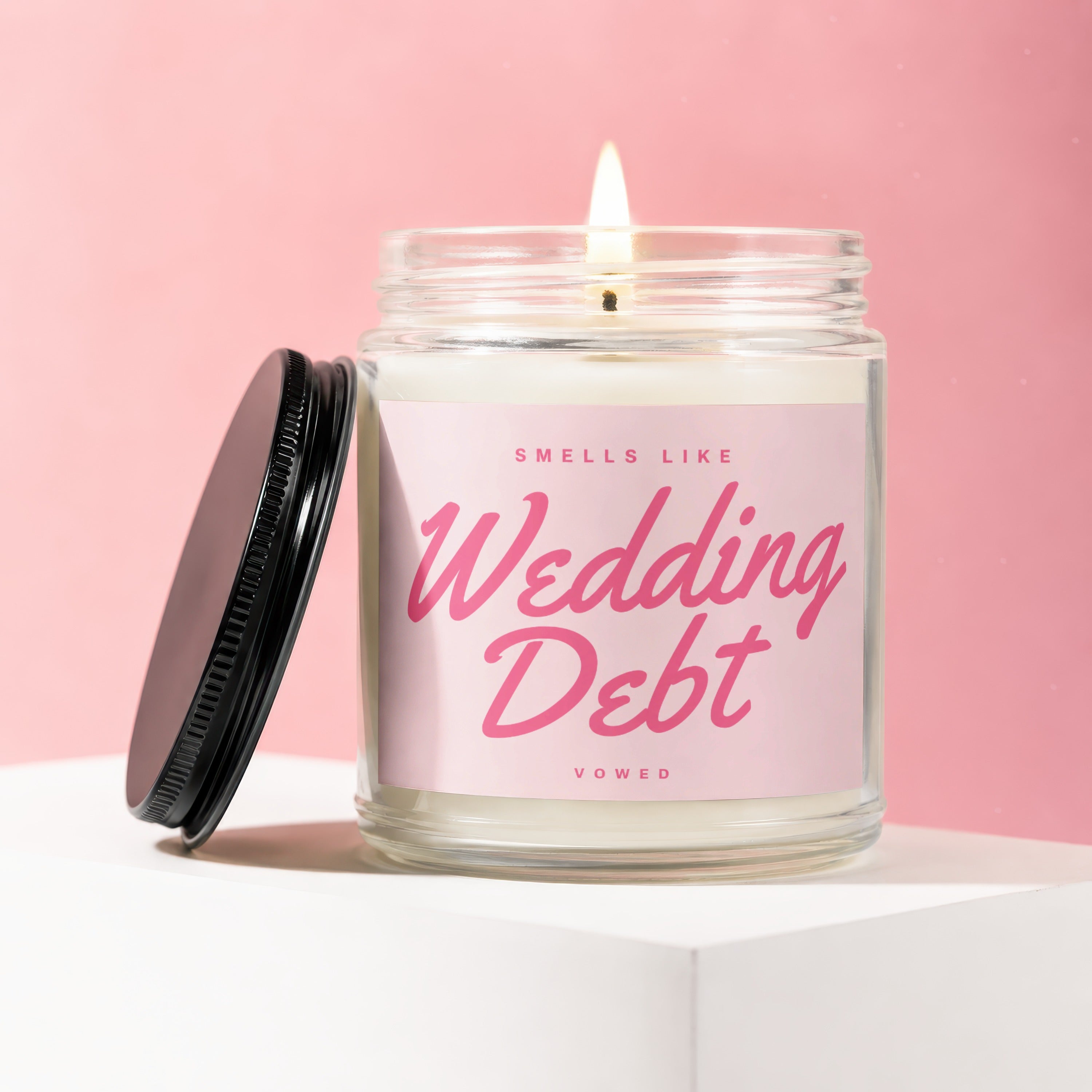 Glass jar candle with pink label reading "Smells Like Wedding Debt Vowed".