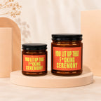 Two amber glass candles with black lids and orange labels reading "You Lit Up That F*cking Ceremony."