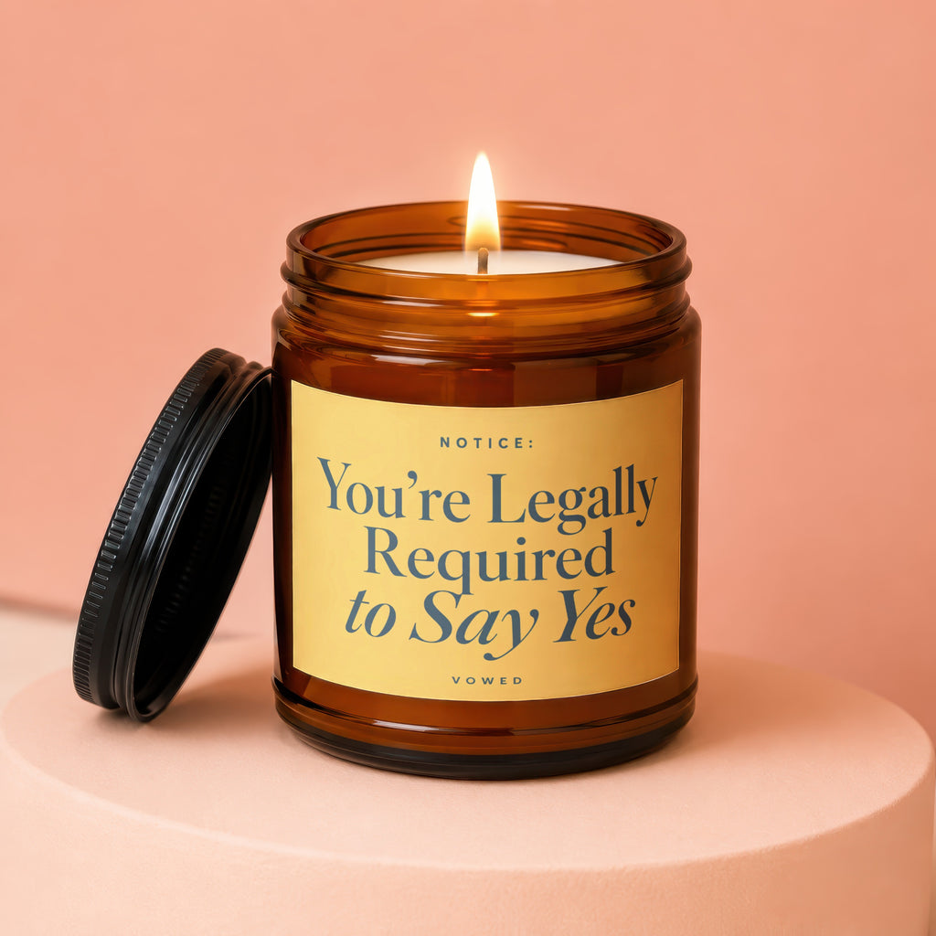 Brown glass candle with lighted wick and label reading "NOTICE: You're Legally Required to Say Yes VOWED."