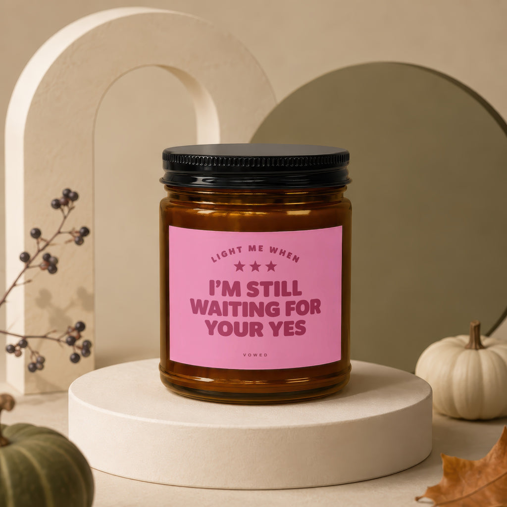 Brown glass candle with pink label reading "LIGHT ME WHEN I'M STILL WAITING FOR YOUR YES".