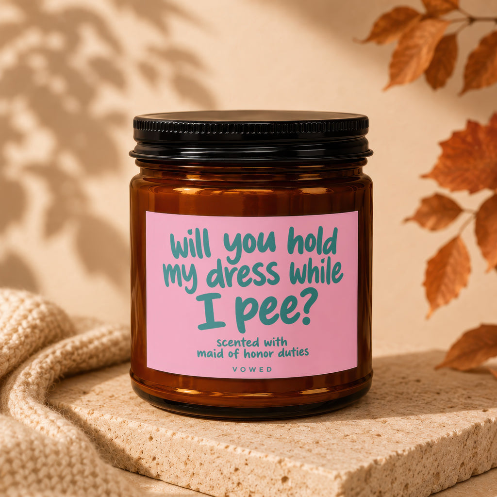 Amber candle with pink label that reads "will you hold my dress while I pee?"