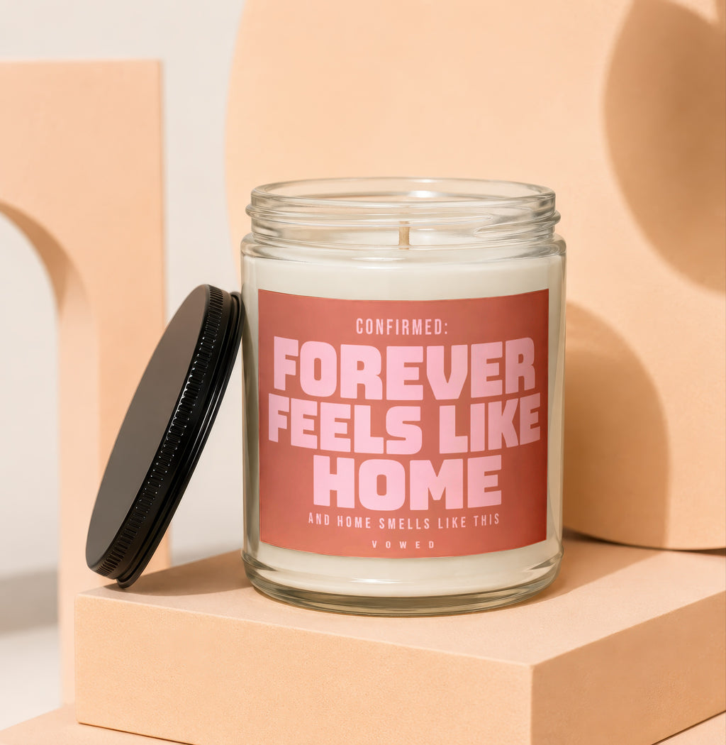 Glass candle with pink label that reads "CONFIRMED: FOREVER FEELS LIKE HOME".