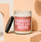 Glass candle with pink label that reads "CONFIRMED: FOREVER FEELS LIKE HOME".