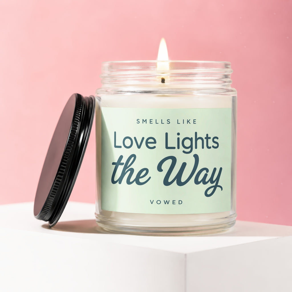 Glass jar candle with lit flame and black lid, reads "Smells Like Love Lights the Way Vowed".