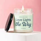 Glass jar candle with lit flame and black lid, reads "Smells Like Love Lights the Way Vowed".