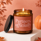 Amber candle with lit wick and lid off, featuring text "Partners, officially".