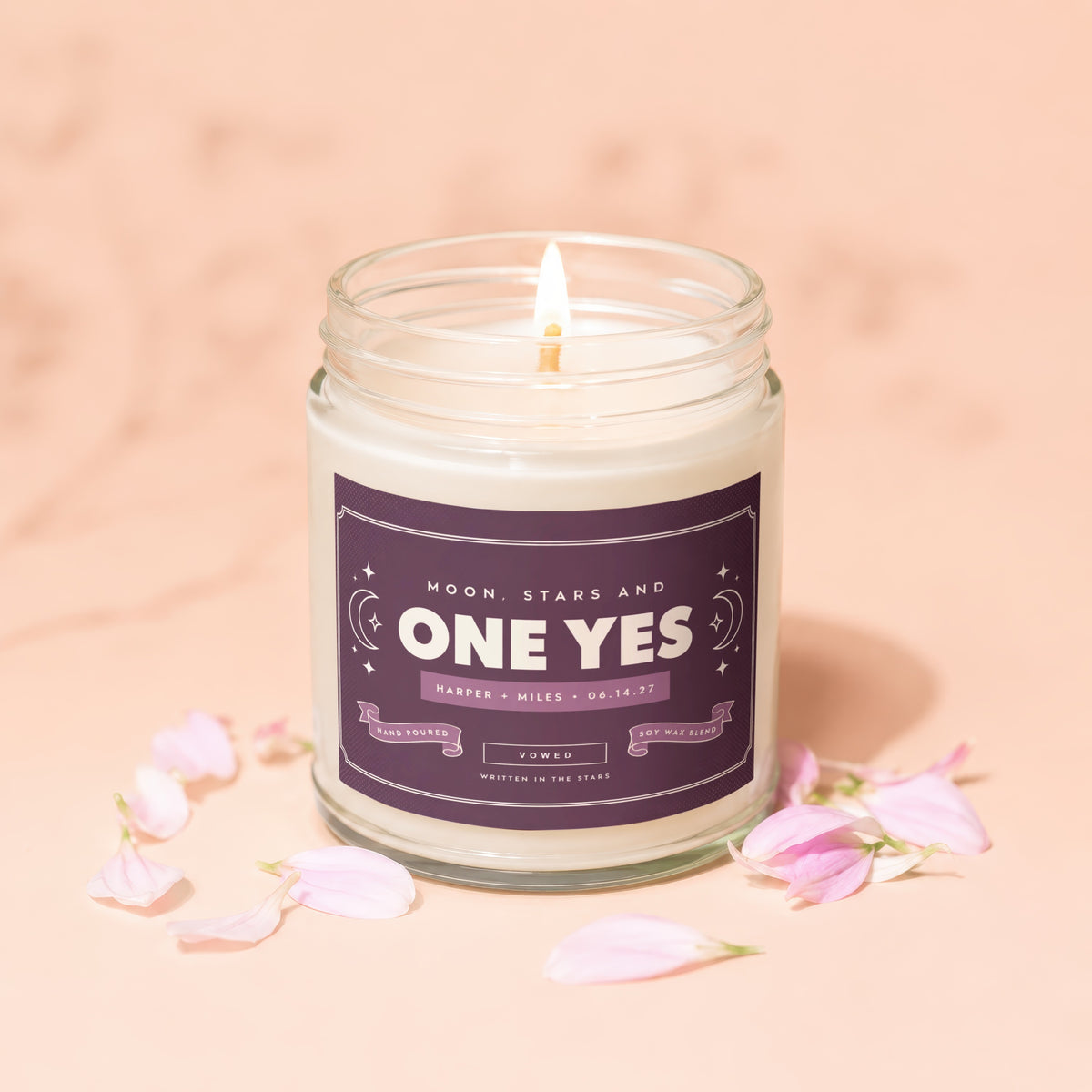 A lit soy wax candle in a clear glass jar with a purple personalized label, surrounded by scattered pink petals.