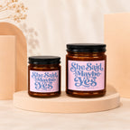 Two amber glass candles with pink labels reading "She Said Maybe Yes" and "And She Meant It".
