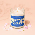 Glass jar candle with blue label reading "HERE'S TO FOREVER" surrounded by pink petals.