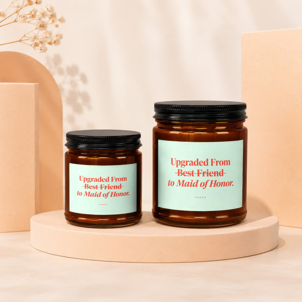 Two amber glass candles with black lids, featuring labels that read "Upgraded From Best Friend to Maid of Honor."