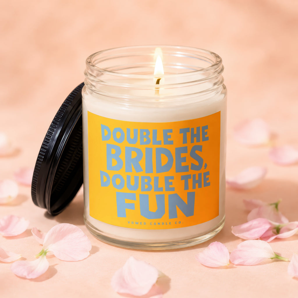 Glass candle with burning wick and black lid, label reads "DOUBLE THE BRIDES, DOUBLE THE FUN."