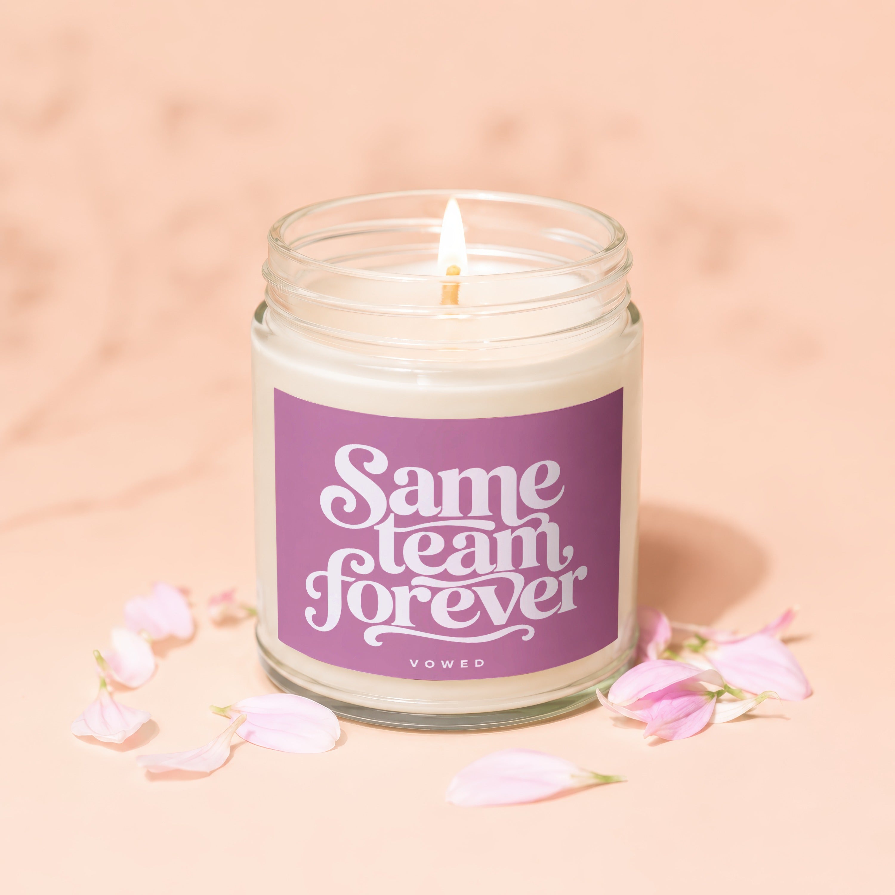 Lit candle with "Same team forever" text on a purple label and pink petals.