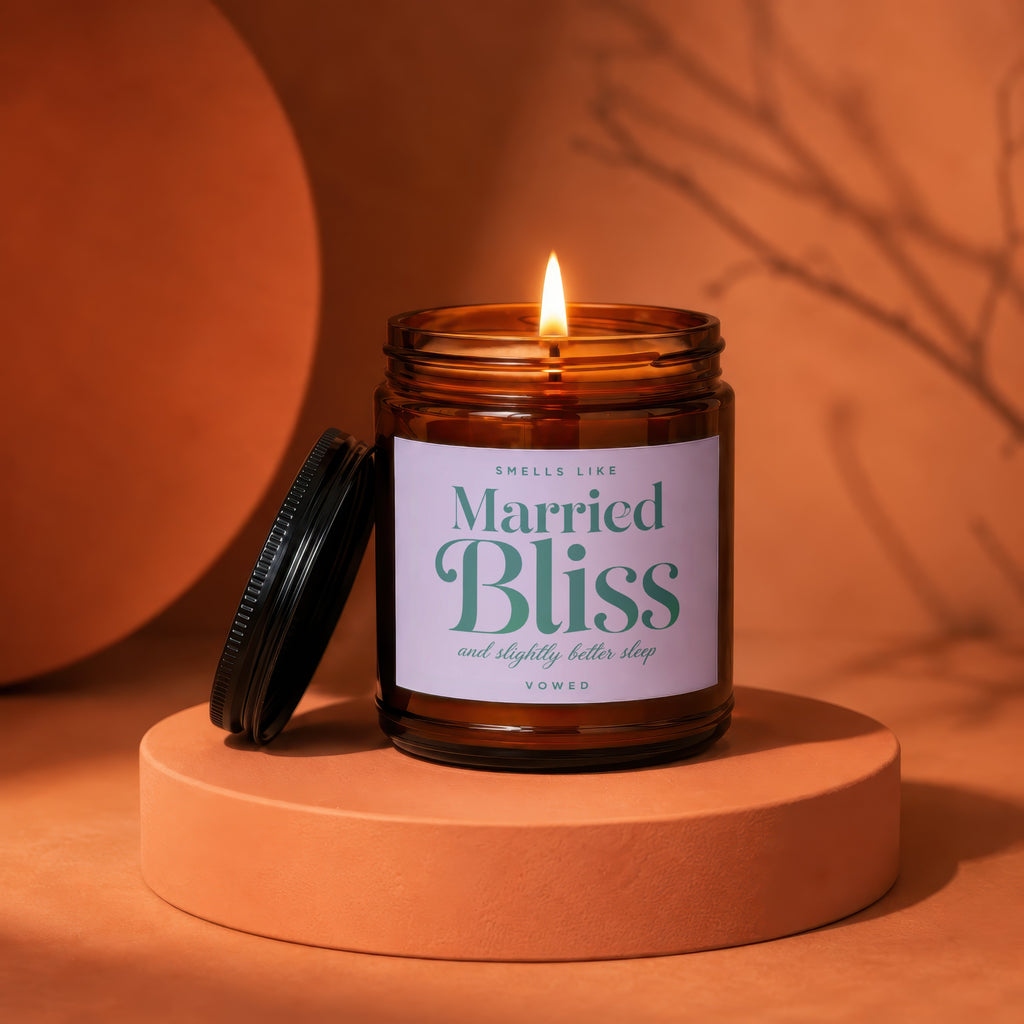 Brown glass candle with lit wick, labeled "Married Bliss and slightly better sleep", on orange surface.