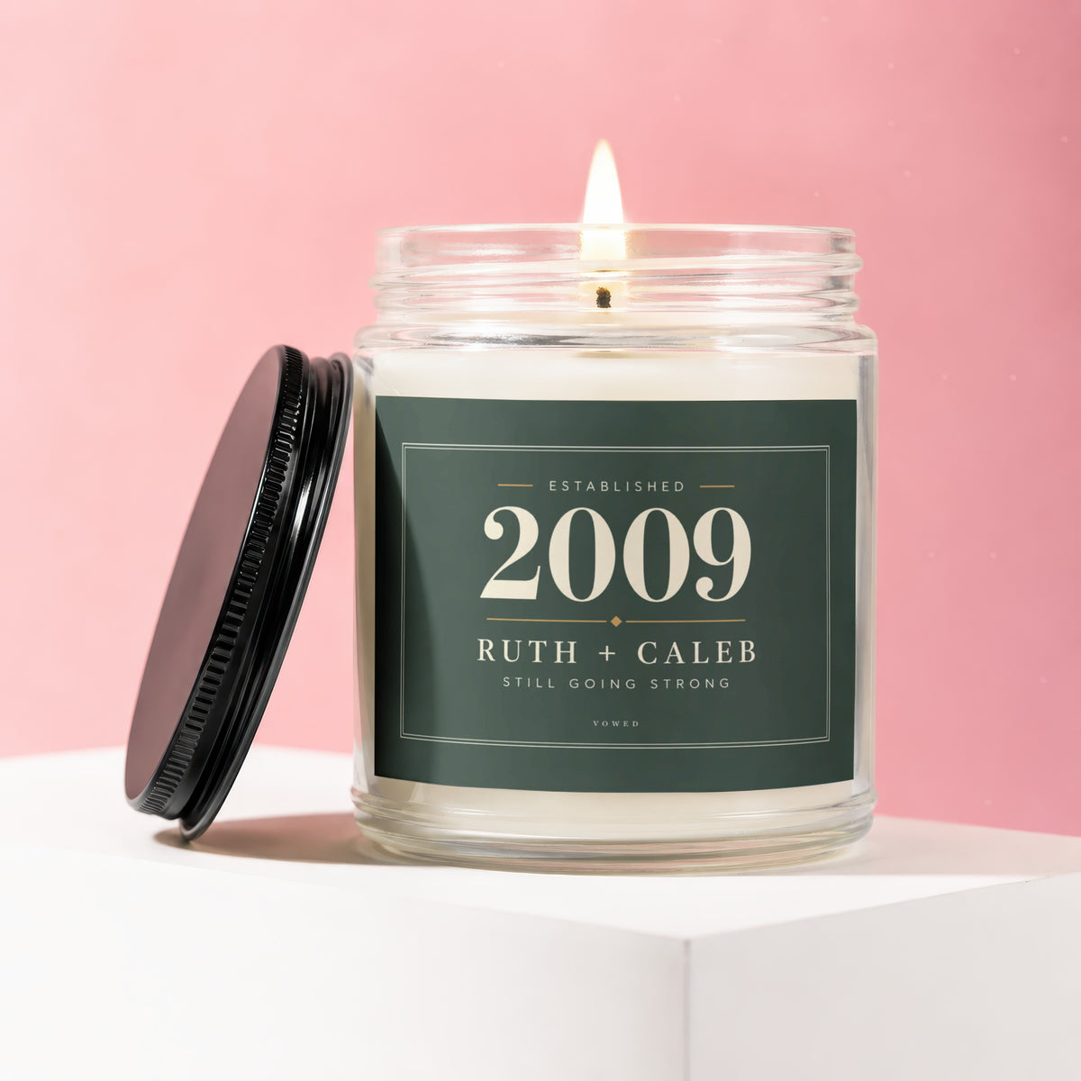 Glass jar candle with lid off, lit wick, and "Established 2009 Ruth + Caleb Still Going Strong" label.
