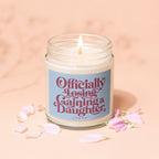 Candle with blue label reading "Officially Losing Gaining a Daughter" with petals.