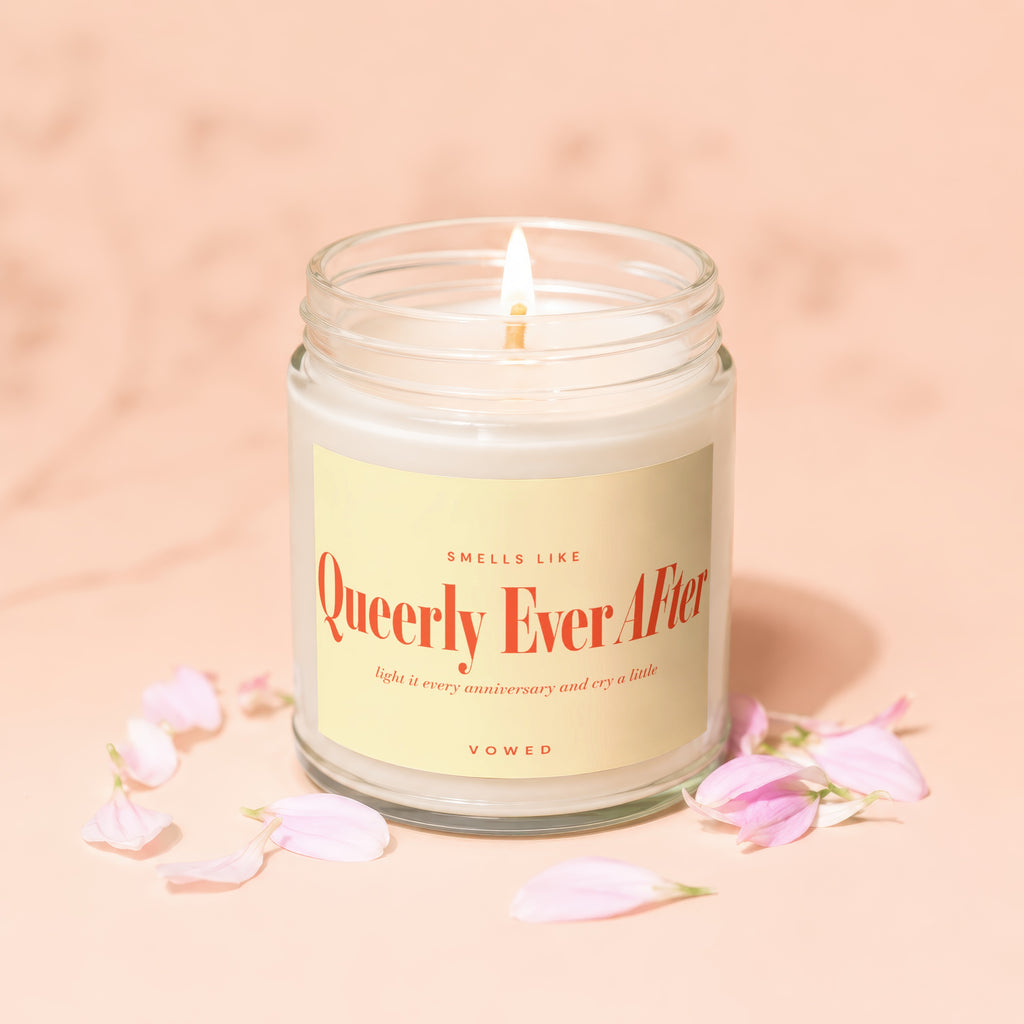 Glass candle with lit wick, yellow label reads "Queerly Ever After", pink flower petals scattered.